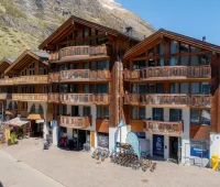 Drei miteinander verbundene, hölzerne Gebäude im Chalet-Stil mit mehreren Balkonen, Geschäften und Fahrrädern, die draußen geparkt sind, vor einer Bergkulisse an einem sonnigen Tag.